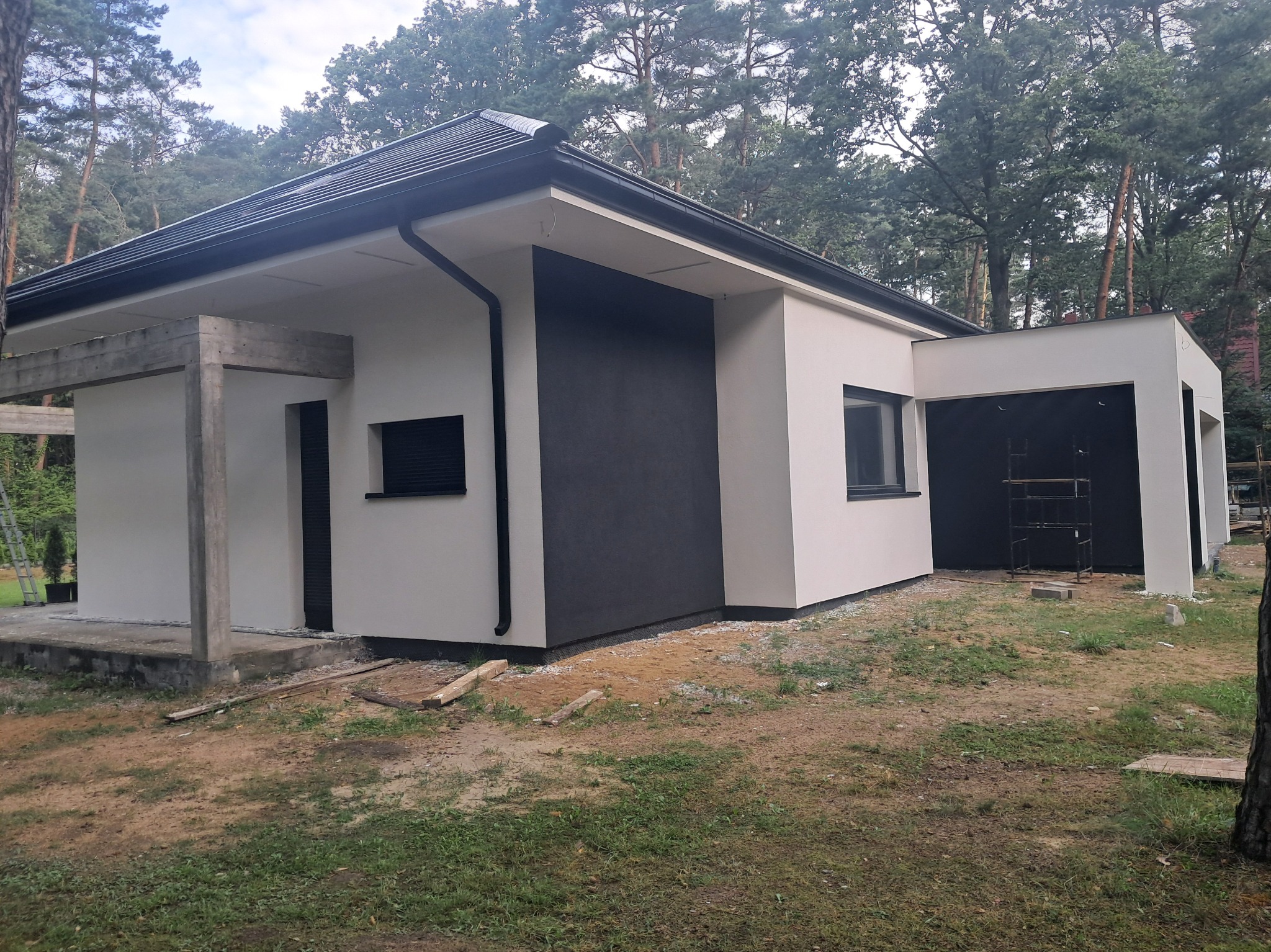 Nowoczesny dom z elewacją w odcieniach bieli i grafitu, otoczony drzewami. Fragment budynku w trakcie wykończenia z widocznymi elementami konstrukcyjnymi.