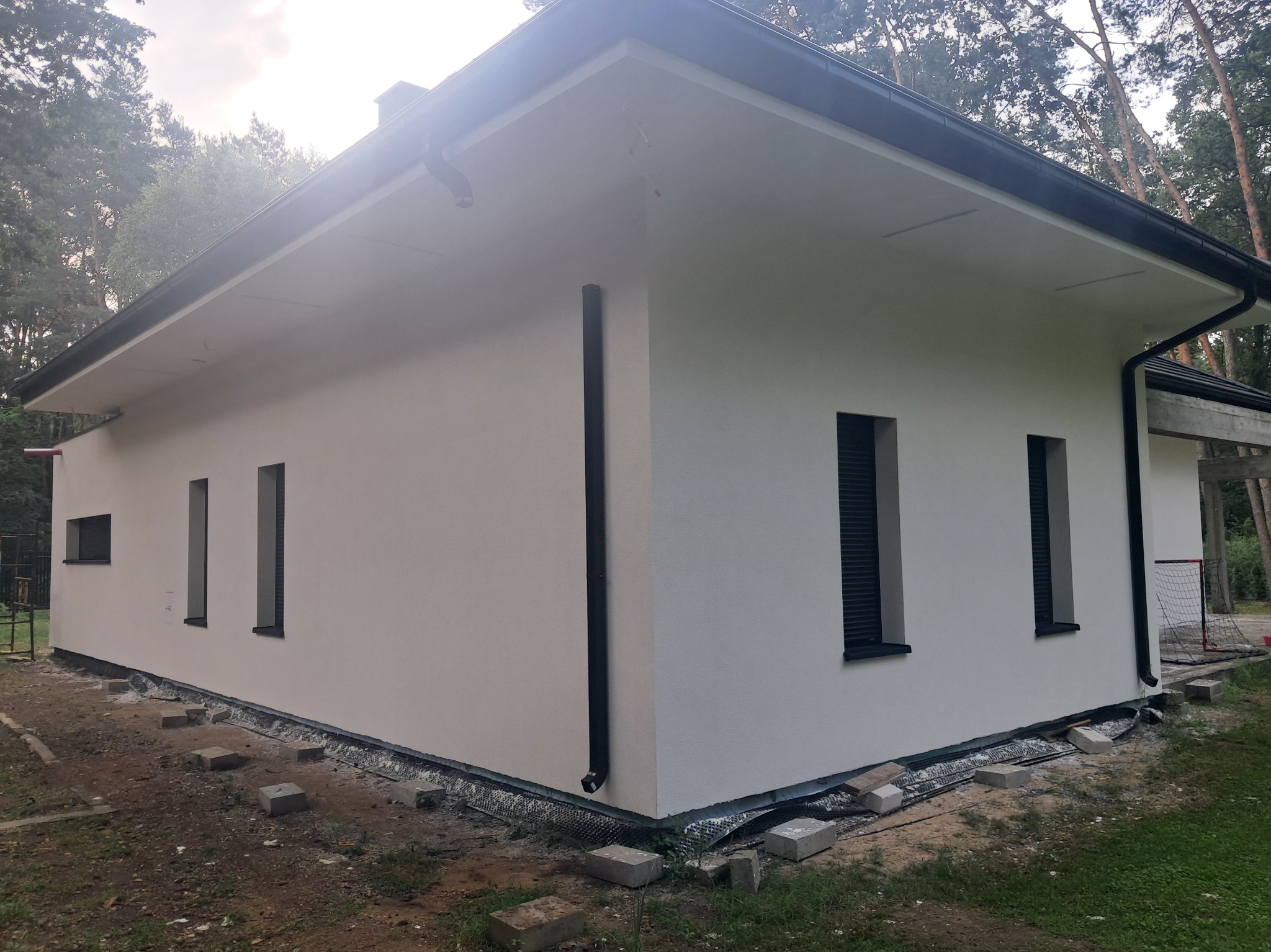 Biały, parterowy dom z czarnymi akcentami (rynny, okna). Wykończona elewacja z widoczną izolacją fundamentów i elementami krajobrazu. Dom otoczony drzewami.