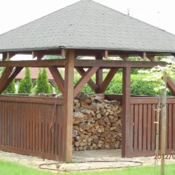Drewniana altana z szarym dachem krytym gontem, wypełniona ułożonym drewnem opałowym, otoczona zielenią ogrodu.