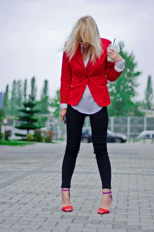 Kobieta w czerwonej marynarce, czarnych spodniach i czerwonych sandałach na obcasie z fioletowymi paskami, trzymająca napój w kubku, stojąca na chodniku.