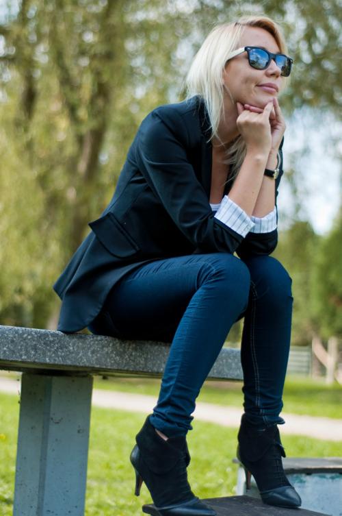 Kobieta w czarnej marynarce, dżinsach i botkach na obcasie siedzi na betonowym murku, opierając brodę na dłoniach i patrząc w dal, ma na sobie białe okulary przeciwsłoneczne.
