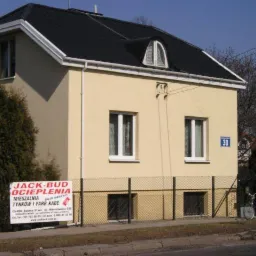 Dwukondygnacyjny dom z beżową elewacją, czarnym dachem i białymi ramami okiennymi, widoczny znak firmowy na ogrodzeniu i rowerzysta przejeżdżający obok.