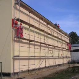Pracownicy na rusztowaniu malują fragment elewacji budynku na czerwono, pozostała część elewacji w kolorze beżowym, widoczne niebo i budynki w tle.
