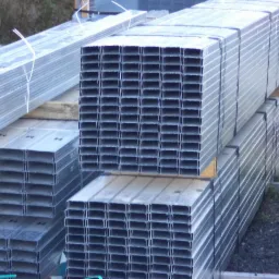 Srebrne profile stalowe do suchej zabudowy, ułożone w paczkach na placu składowym, widoczne wiązania i perforowane przekroje.