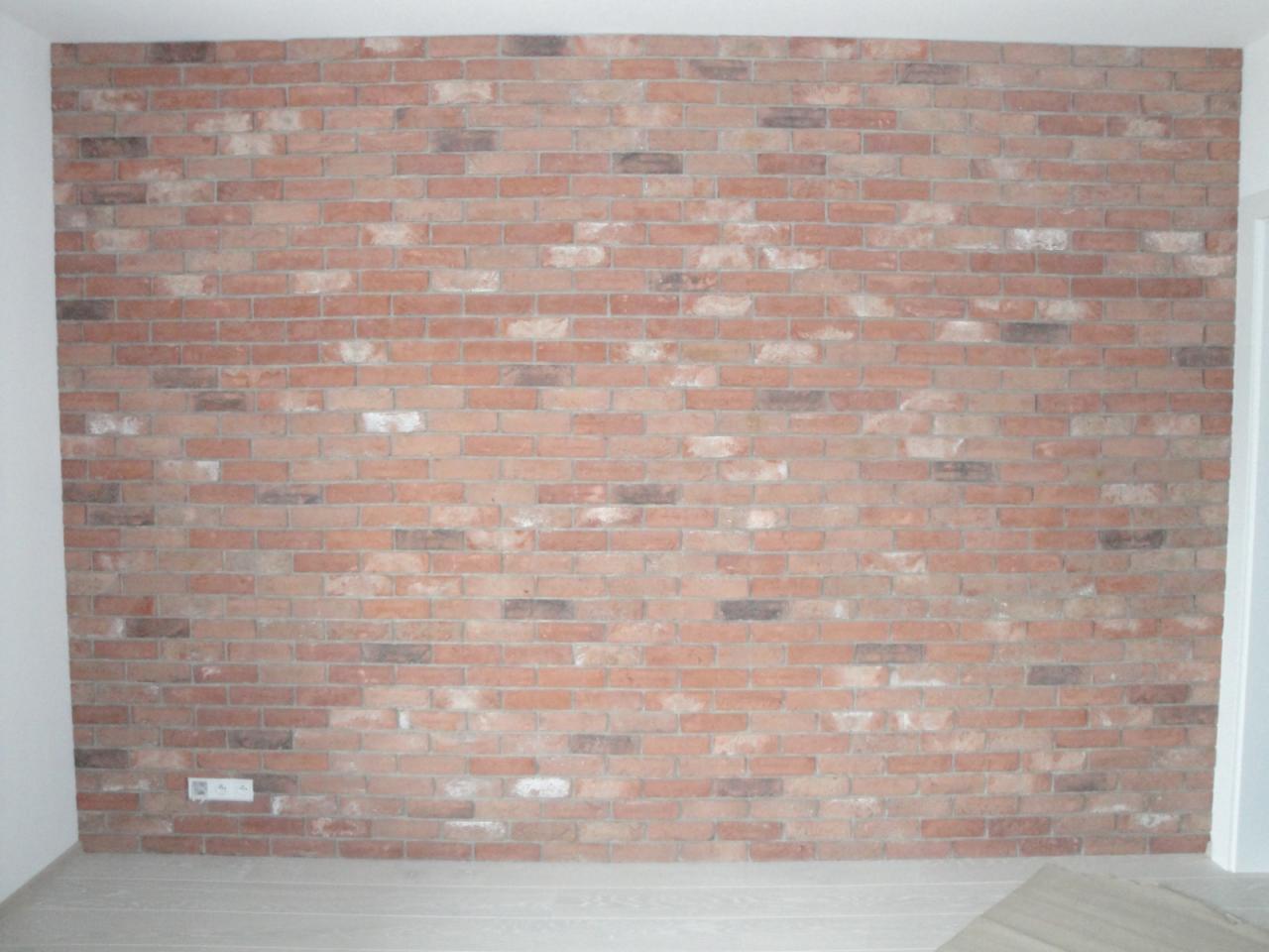 Ściana wykończona czerwoną cegłą w stylu retro z widocznym gniazdkiem elektrycznym i fragmentem drewnianej podłogi.