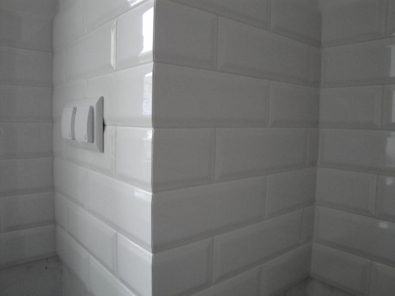 Białe, błyszczące płytki ułożone w cegiełkę na ścianie, widoczny narożnik oraz gniazdko elektryczne z dwoma włącznikami.