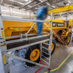 VOLVO MASZYNY BUDOWLANE POLSKA Sp. z o.o. - Żółta wywrotka Volvo A30G w hali produkcyjnej, spawanie konstrukcji z iskrami, drabina aluminiowa z boku, widoczny suwnica i wentylacja przemysłowa.