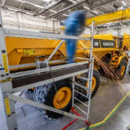 Żółta wywrotka Volvo A30G w hali produkcyjnej, spawanie konstrukcji z iskrami, drabina aluminiowa z boku, widoczny suwnica i wentylacja przemysłowa.