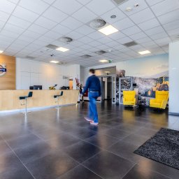 VOLVO MASZYNY BUDOWLANE POLSKA Sp. z o.o. - Wnętrze salonu Volvo z recepcją, logiem firmy, muralem z koparką i żółtymi fotelami, z przechodzącą osobą.