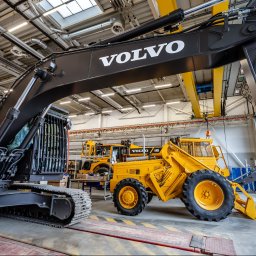 VOLVO MASZYNY BUDOWLANE POLSKA Sp. z o.o. - Wnętrze hali produkcyjnej w Młochowie z nową, czarną koparką gąsienicową Volvo na pierwszym planie, obok żółta ładowarka kołowa Volvo A30G i inne maszyny w tle, oświetlenie sufitowe.