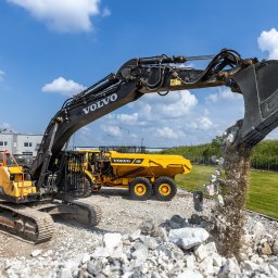 VOLVO MASZYNY BUDOWLANE POLSKA Sp. z o.o. - Żółta koparka Volvo EC380E i wywrotka A30G na żwirowym placu, z łyżką koparki wysypującą gruz, w tle budynek i błękitne niebo z chmurami.