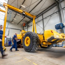 VOLVO MASZYNY BUDOWLANE POLSKA Sp. z o.o. - Żółta maszyna budowlana pod suwnicą w hali serwisowej, dwóch mechaników w ruchu, widoczne ślady użytkowania na maszynie.