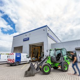 VOLVO MASZYNY BUDOWLANE POLSKA Sp. z o.o. - Zielona ładowarka Volvo z łyżką na tle budynku serwisu Volvo i dwóch busów serwisowych Volvo, z przechodzącym mężczyzną. Ładowarka stoi na kostce brukowej.