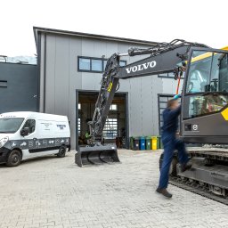 VOLVO MASZYNY BUDOWLANE POLSKA Sp. z o.o. - Pracownik w kombinezonie wchodzi do kabiny nowej, żółto-czarnej koparki Volvo na gąsienicach, z przodu biuro serwisu z zaparkowanymi busami serwisowymi Volvo