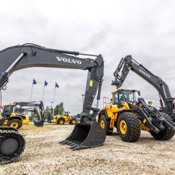 VOLVO MASZYNY BUDOWLANE POLSKA Sp. z o.o. - Grupa nowych, żółto-czarnych maszyn budowlanych Volvo, w tym koparka gąsienicowa EC350EL, ładowarka kołowa i inne, stojących na placu z flagami w tle.