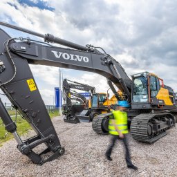 VOLVO MASZYNY BUDOWLANE POLSKA Sp. z o.o. - Rząd nowych, szarych i żółtych koparek Volvo na żwirowym placu, z rozmytą sylwetką osoby w kamizelce odblaskowej przechodzącej przed największą z nich.