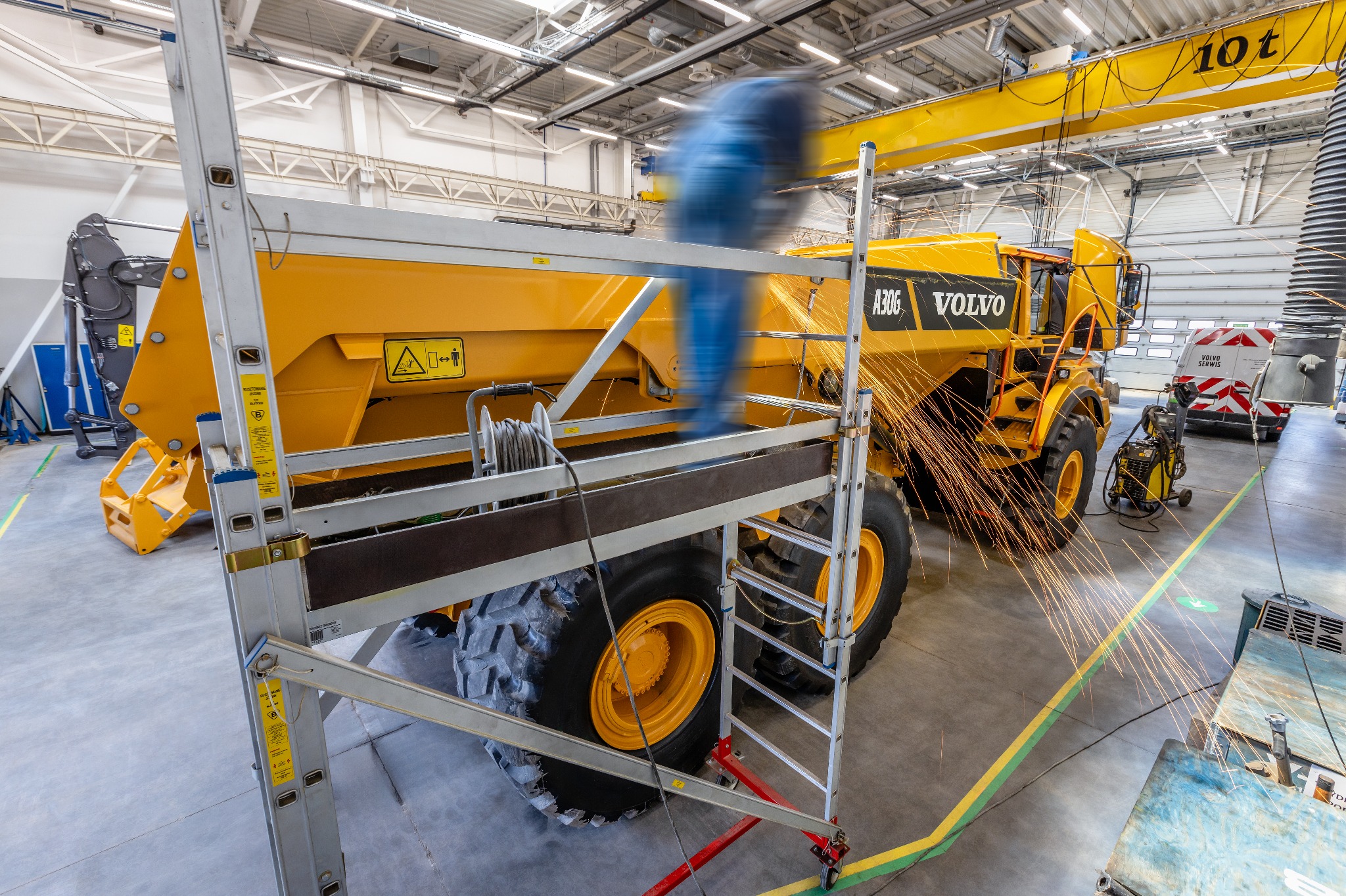 Żółta wywrotka Volvo A30G w hali produkcyjnej, spawanie konstrukcji z iskrami, drabina aluminiowa z boku, widoczny suwnica i wentylacja przemysłowa.