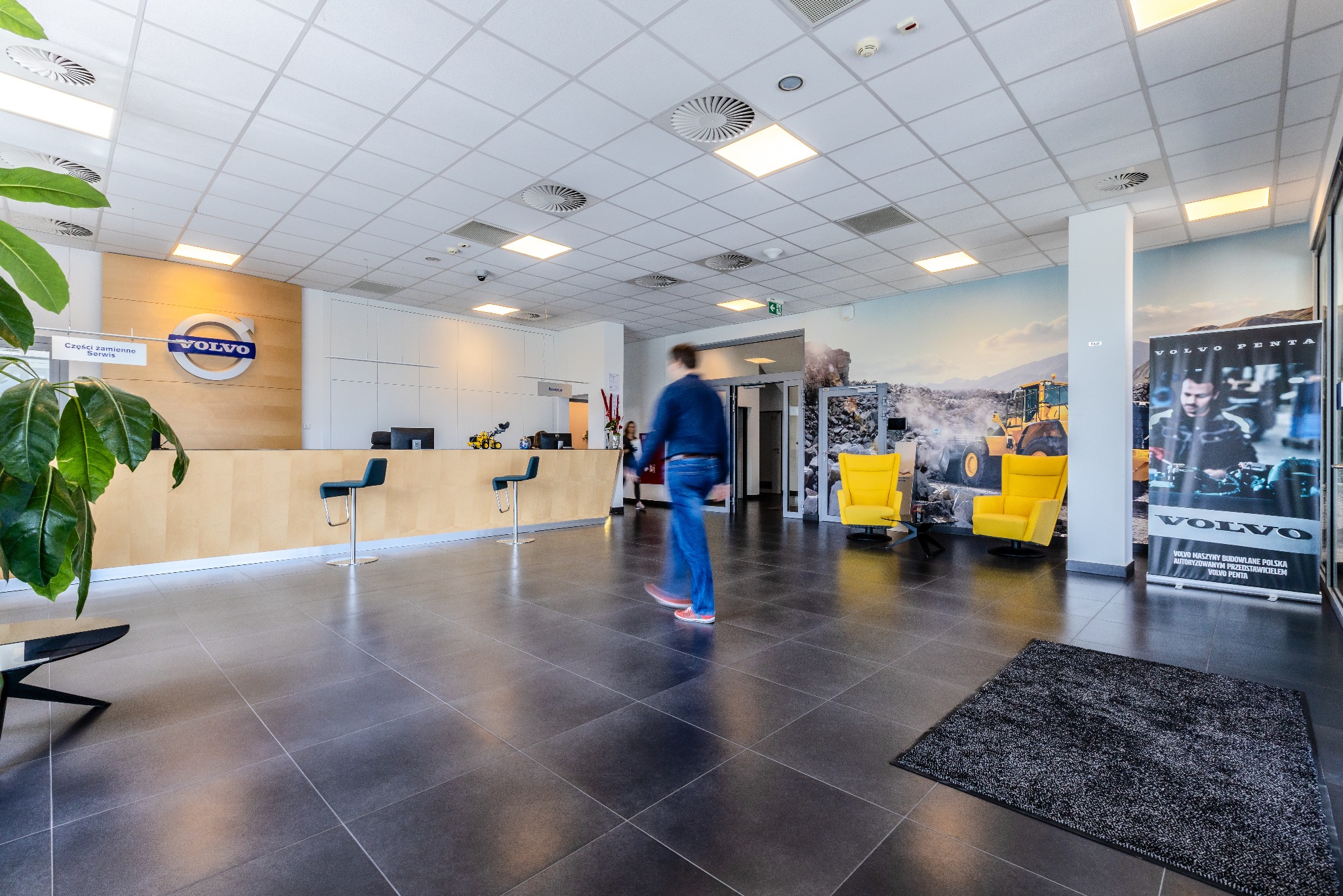 Wnętrze salonu Volvo z recepcją, logiem firmy, muralem z koparką i żółtymi fotelami, z przechodzącą osobą.