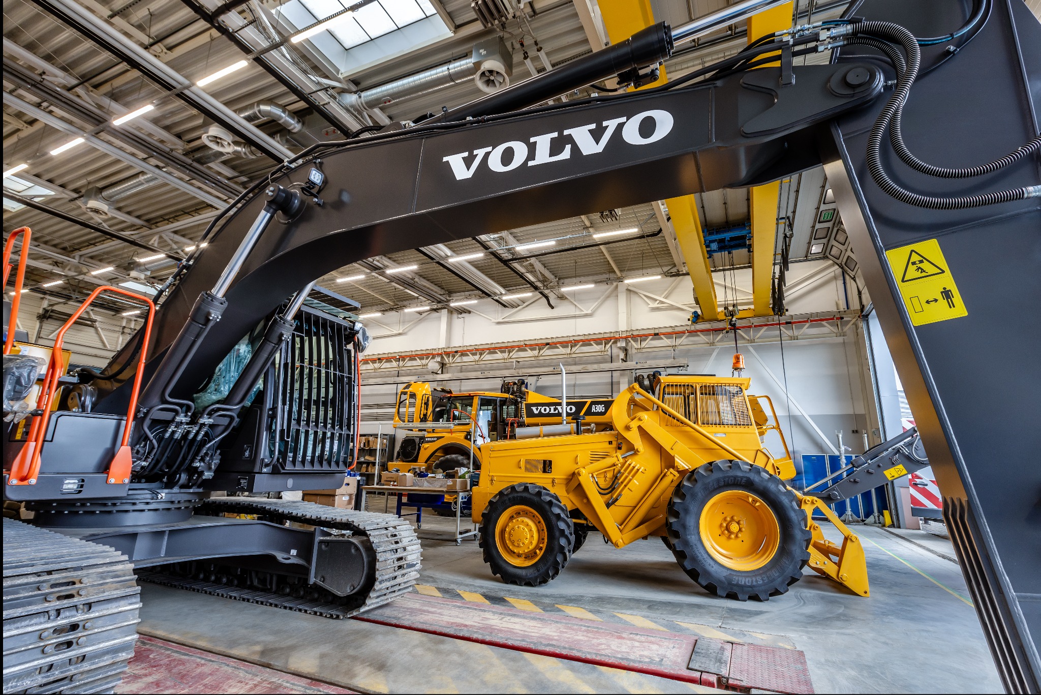Wnętrze hali produkcyjnej w Młochowie z nową, czarną koparką gąsienicową Volvo na pierwszym planie, obok żółta ładowarka kołowa Volvo A30G i inne maszyny w tle, oświetlenie sufitowe.