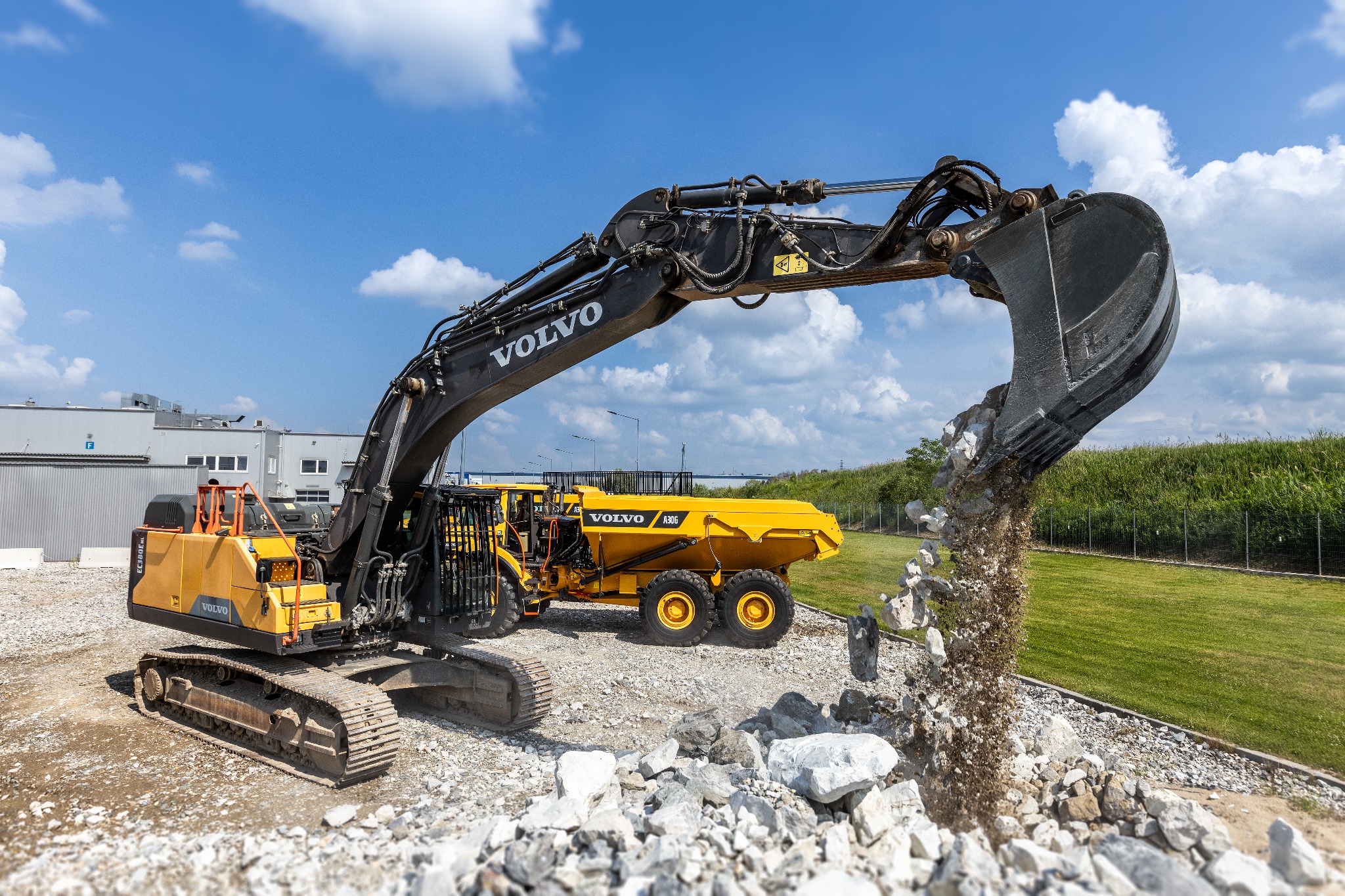 Żółta koparka Volvo EC380E i wywrotka A30G na żwirowym placu, z łyżką koparki wysypującą gruz, w tle budynek i błękitne niebo z chmurami.