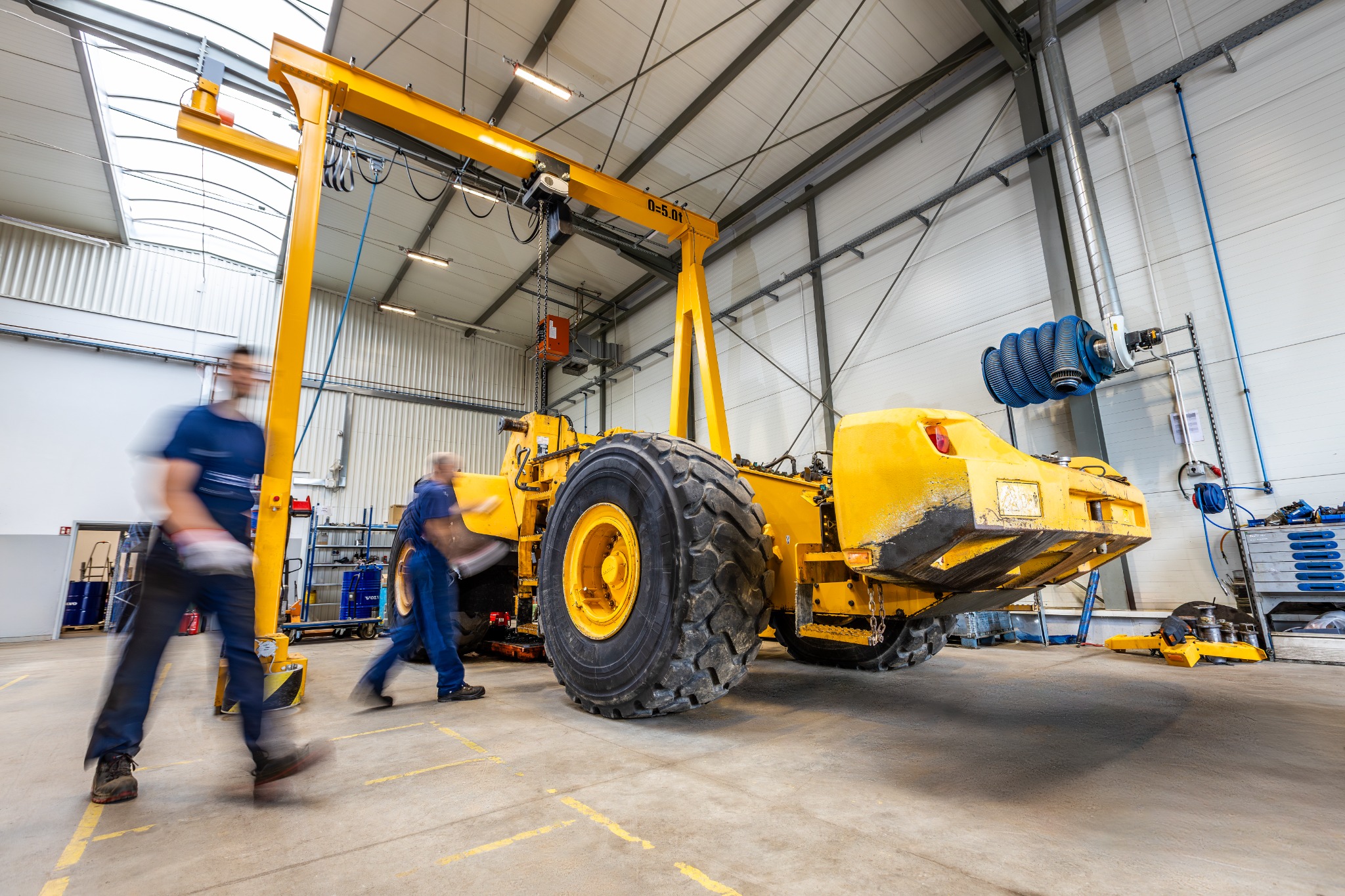 Żółta maszyna budowlana pod suwnicą w hali serwisowej, dwóch mechaników w ruchu, widoczne ślady użytkowania na maszynie.