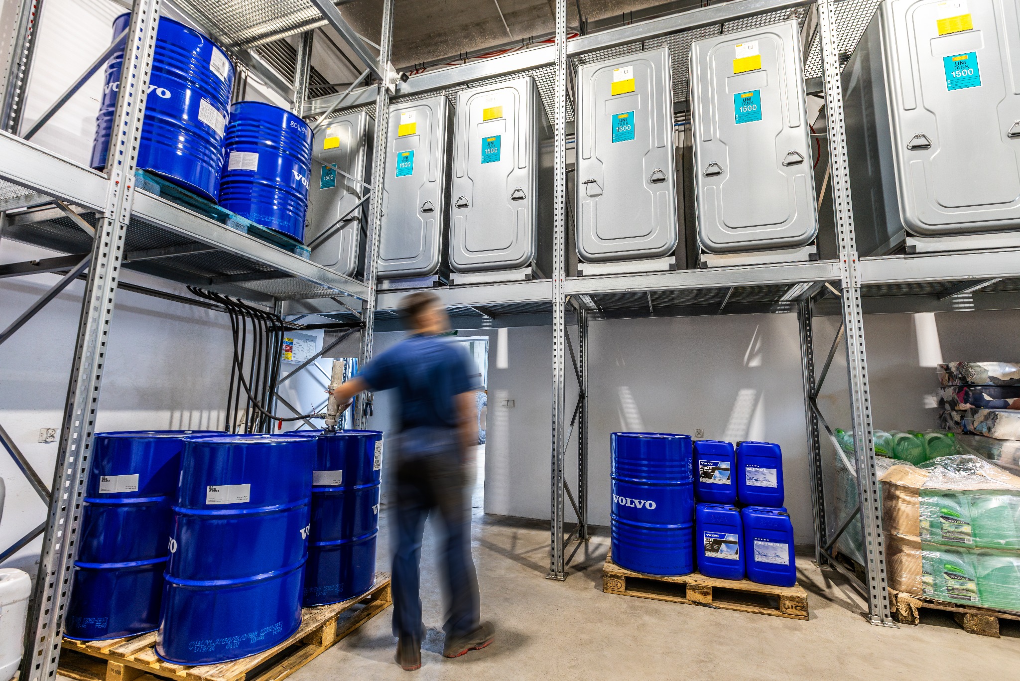 Wnętrze magazynu z wysokimi regałami, na których składowane są niebieskie beczki z logo Volvo oraz szare przenośne toalety UNI TANK 1500, rozmyta sylwetka osoby przy beczkach.