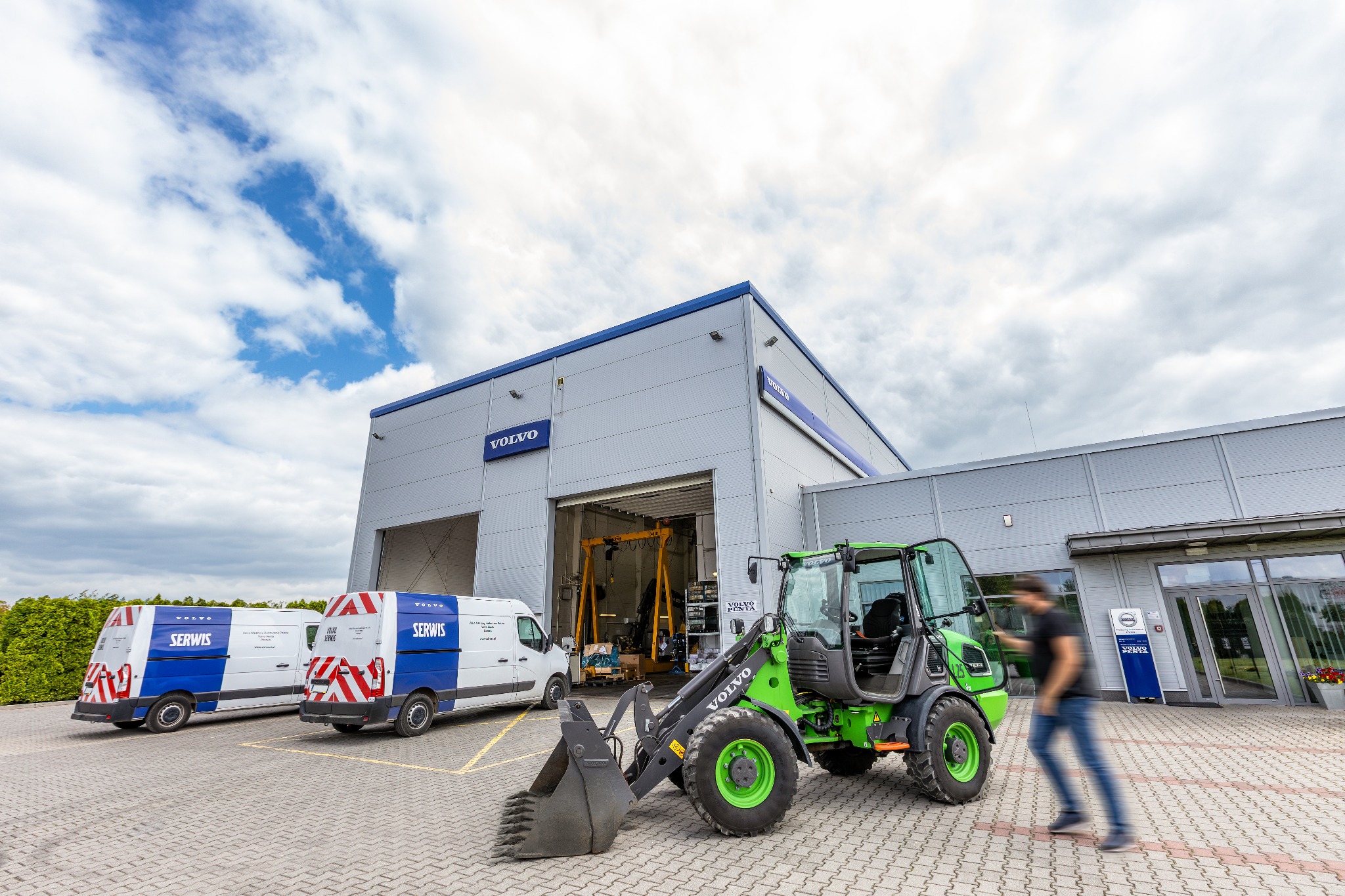 Zielona ładowarka Volvo z łyżką na tle budynku serwisu Volvo i dwóch busów serwisowych Volvo, z przechodzącym mężczyzną. Ładowarka stoi na kostce brukowej.