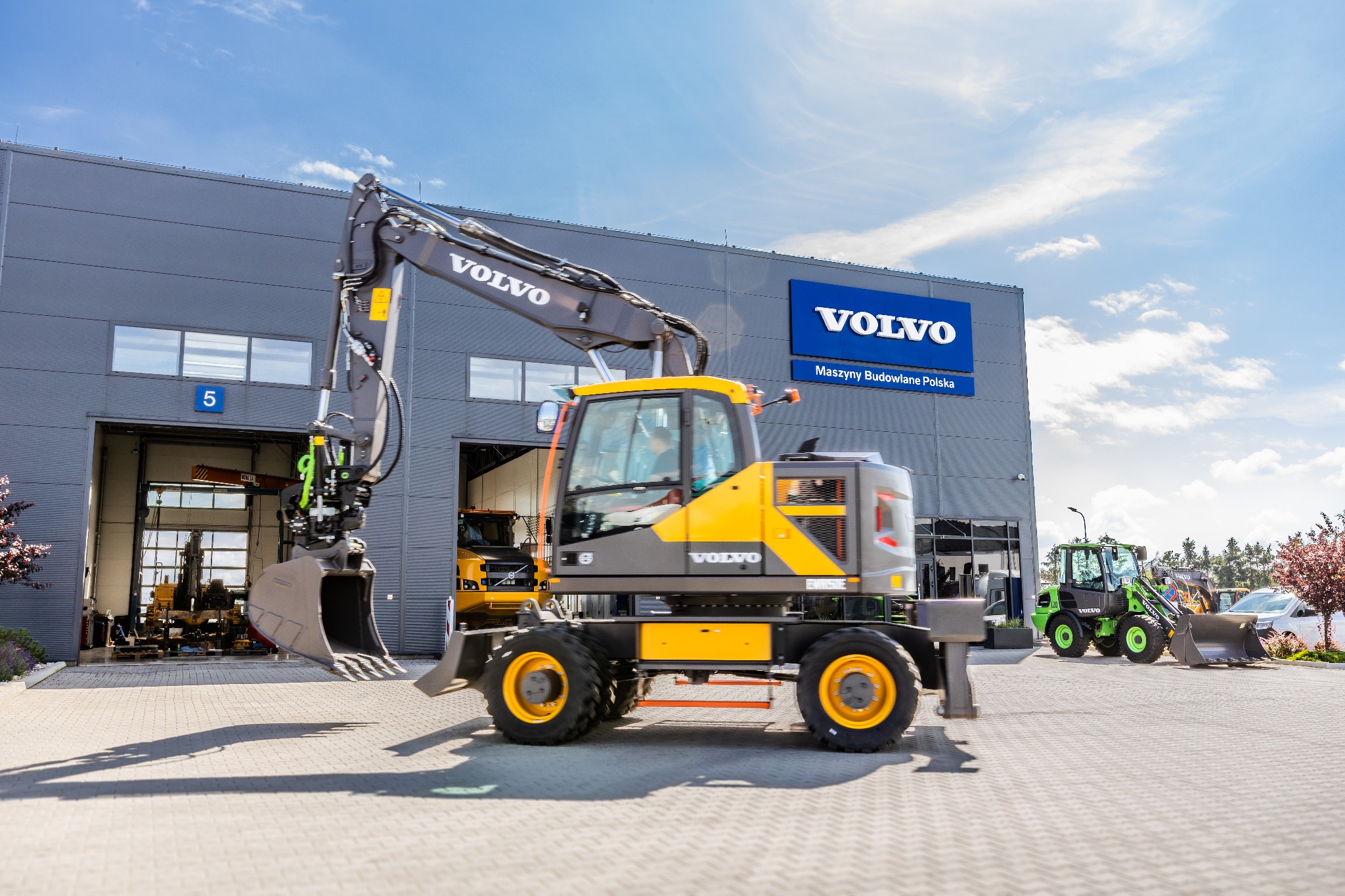 Żółto-szara koparka kołowa marki Volvo na tle budynku z logo Volvo, z widoczną inną maszyną budowlaną w tle.