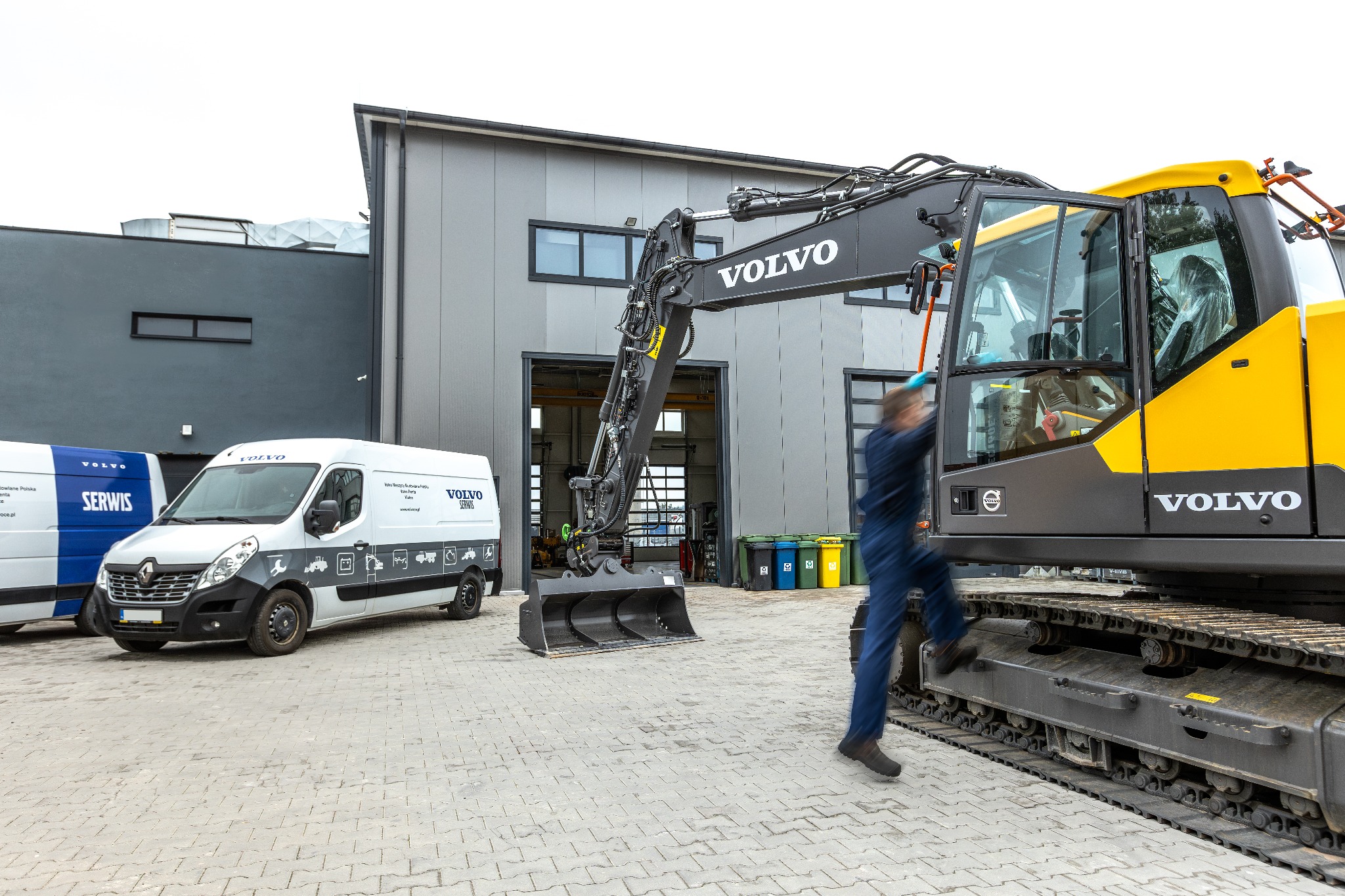 Pracownik w kombinezonie wchodzi do kabiny nowej, żółto-czarnej koparki Volvo na gąsienicach, z przodu biuro serwisu z zaparkowanymi busami serwisowymi Volvo