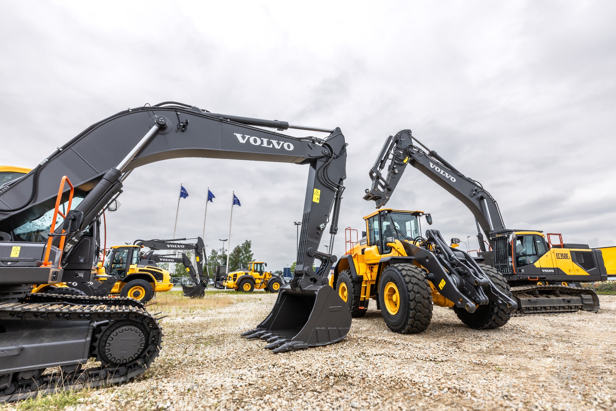 Grupa nowych, żółto-czarnych maszyn budowlanych Volvo, w tym koparka gąsienicowa EC350EL, ładowarka kołowa i inne, stojących na placu z flagami w tle.