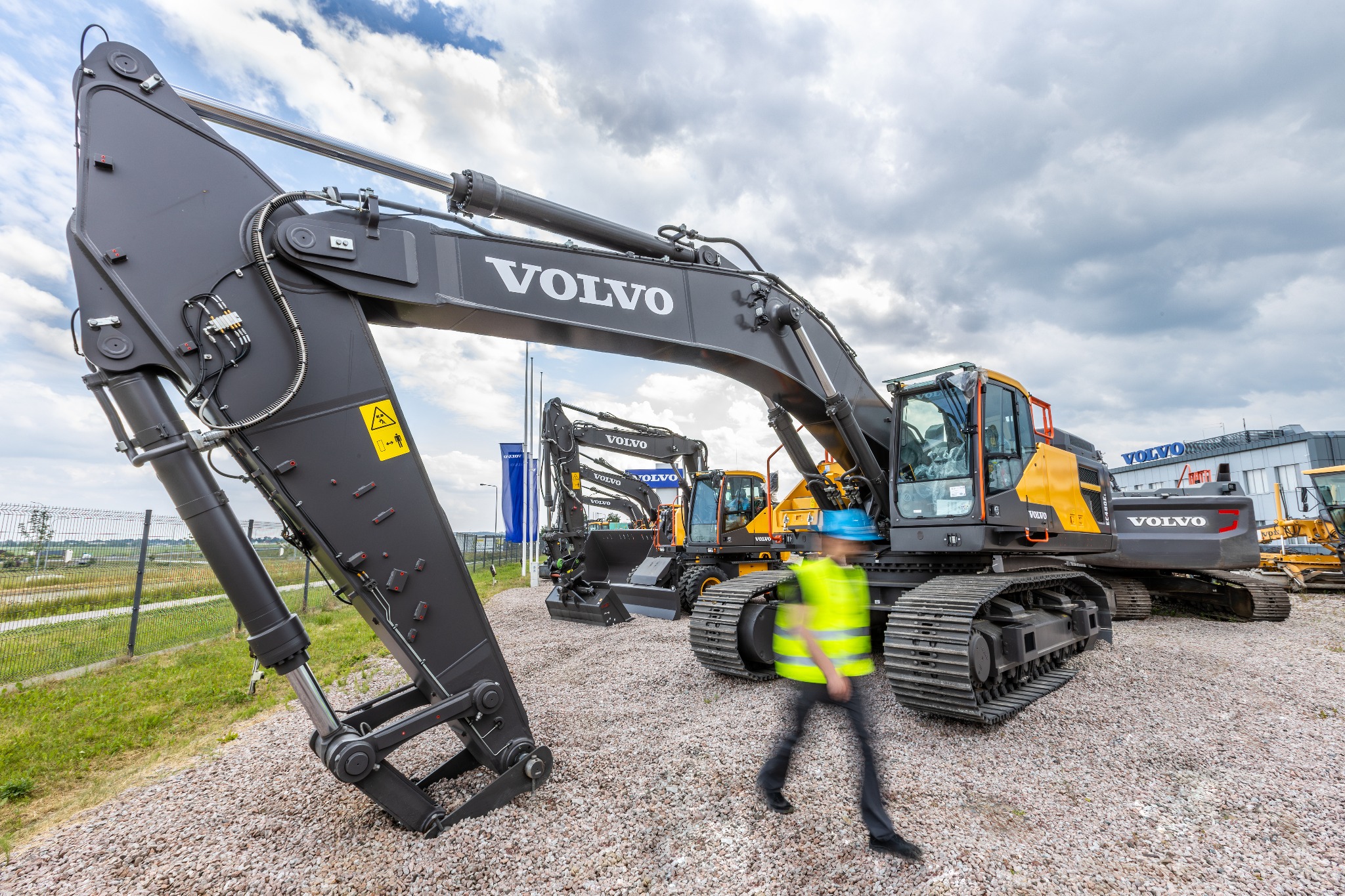 Rząd nowych, szarych i żółtych koparek Volvo na żwirowym placu, z rozmytą sylwetką osoby w kamizelce odblaskowej przechodzącej przed największą z nich.