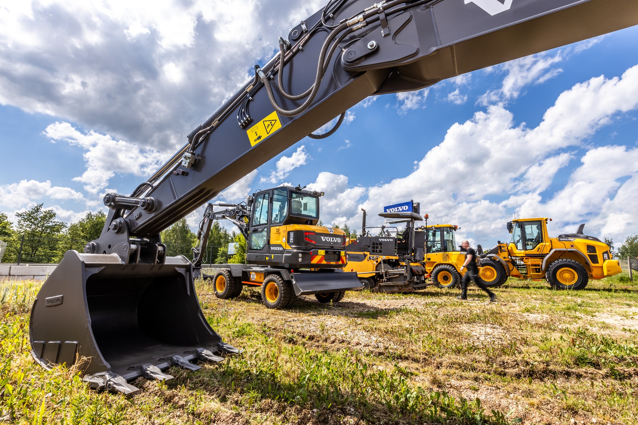 Żółta koparka kołowa Volvo EW160E z widocznym łyżką na pierwszym planie, w tle inne maszyny budowlane i osoba przechodząca obok.