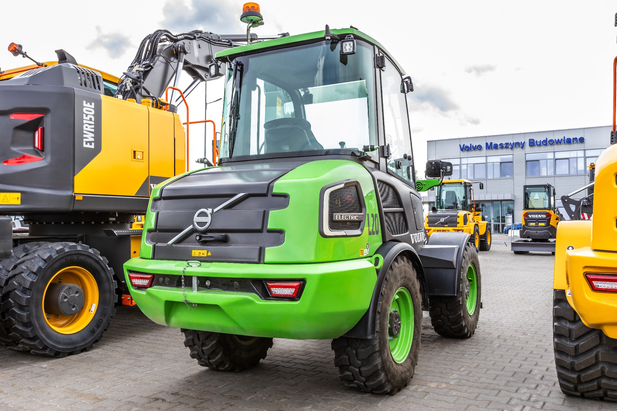 Nowoczesna, jasnozielona ładowarka kołowa Volvo L20 Electric na placu z kostki brukowej, z żółtą koparką kołową EWR150E po lewej i budynkiem Volvo Maszyny Budowlane w tle.