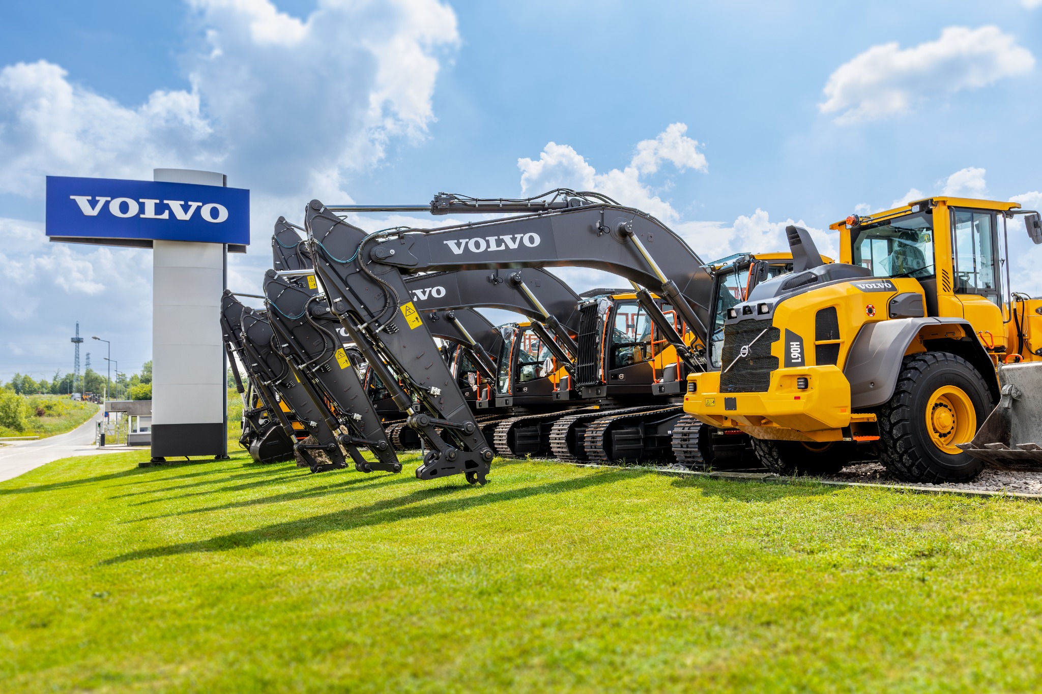 Rząd nowych, czarnych koparek Volvo i żółta ładowarka Volvo L90H na trawniku obok znaku firmowego Volvo, błękitne niebo z chmurami w tle.