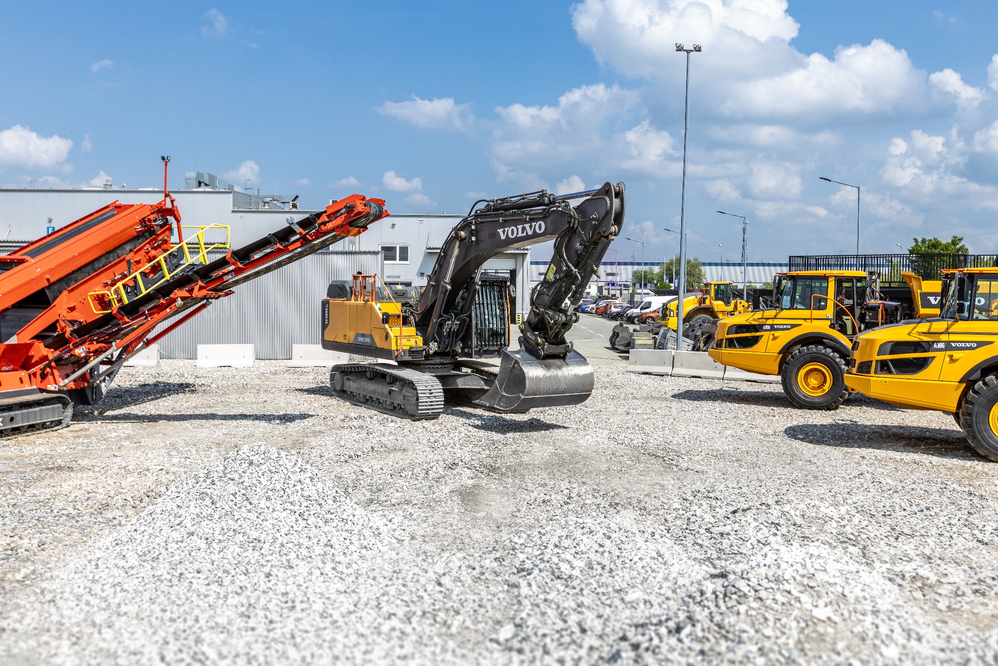 Ciężki sprzęt budowlany Volvo: czarna koparka gąsienicowa, pomarańczowy przesiewacz taśmowy i żółte ładowarki kołowe na placu z kruszywem, w tle budynek i zaparkowane samochody.
