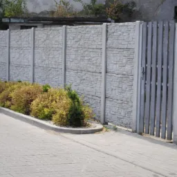 Szary betonowy płot z bramką i roślinnością przy chodniku z kostki brukowej, widoczne słupki i panele imitujące kamień.