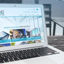 Laptop wyświetlający stronę internetową firmy Ardeno Stolarka prezentującą nowoczesne okna aluminiowe i ściany fasadowe, umieszczony na drewnianym stole na zewnątrz.