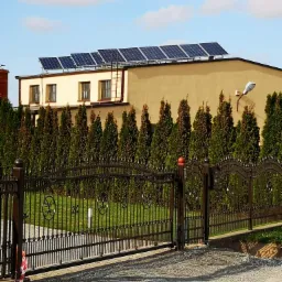Elewacja budynku z panelami słonecznymi na dachu, widoczna za ozdobnym, metalowym ogrodzeniem i żywopłotem z tui.