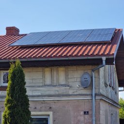 Phu Luxury Marcin Dąbrowski - Panele fotowoltaiczne na dachu domu z czerwoną dachówką, widok z dołu, błękitne niebo w tle. Na ścianie budynku antena satelitarna i numer domu.