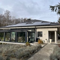 Phu Luxury Marcin Dąbrowski - Dom jednorodzinny z panelami fotowoltaicznymi na dachu, widok z ogrodu. Szara elewacja, przeszklona oranżeria, kostka brukowa i roślinność na pierwszym planie.