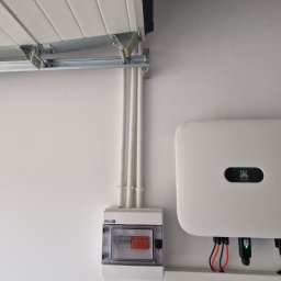 Phu Luxury Marcin Dąbrowski - Biała ściana z zamontowaną skrzynką elektryczną, przewodami i falownikiem Huawei z zielonymi diodami. Widoczne rury instalacyjne i fragment bramy garażowej.