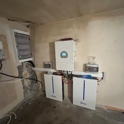 Phu Luxury Marcin Dąbrowski - Białe magazyny energii Deye z inwerterem i rozdzielnicą na ścianie w surowym pomieszczeniu. Widoczne kable i instalacje elektryczne.
