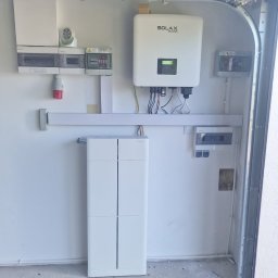 Phu Luxury Marcin Dąbrowski - Biały magazyn energii z falownikiem Solax w garażu, widoczne zabezpieczenia elektryczne i okablowanie. Minimalistyczny design, surowe wykończenie podłogi.