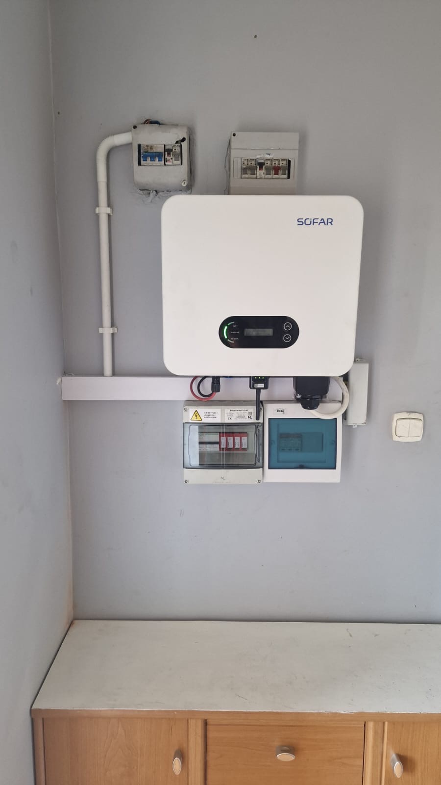 Biały inwerter Sofar zamontowany na szarej ścianie nad drewnianą komodą, z widocznymi przewodami, bezpiecznikami i skrzynkami elektrycznymi. Instalacja fotowoltaiczna wewnątrz budynku.
