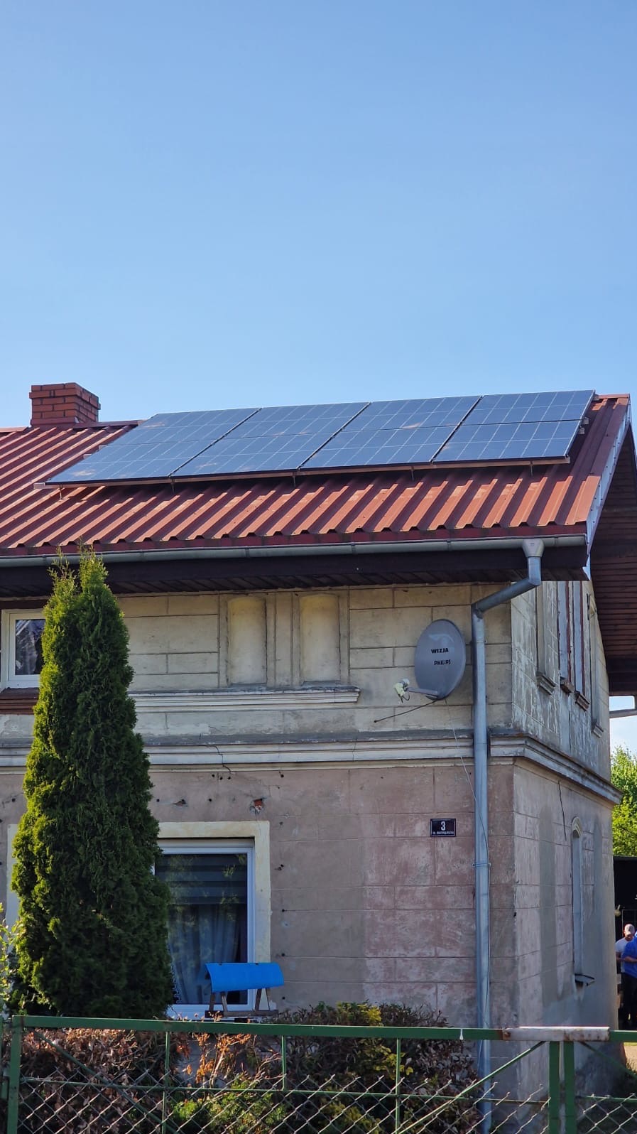Panele fotowoltaiczne na dachu domu z czerwoną dachówką, widok z dołu, błękitne niebo w tle. Na ścianie budynku antena satelitarna i numer domu.
