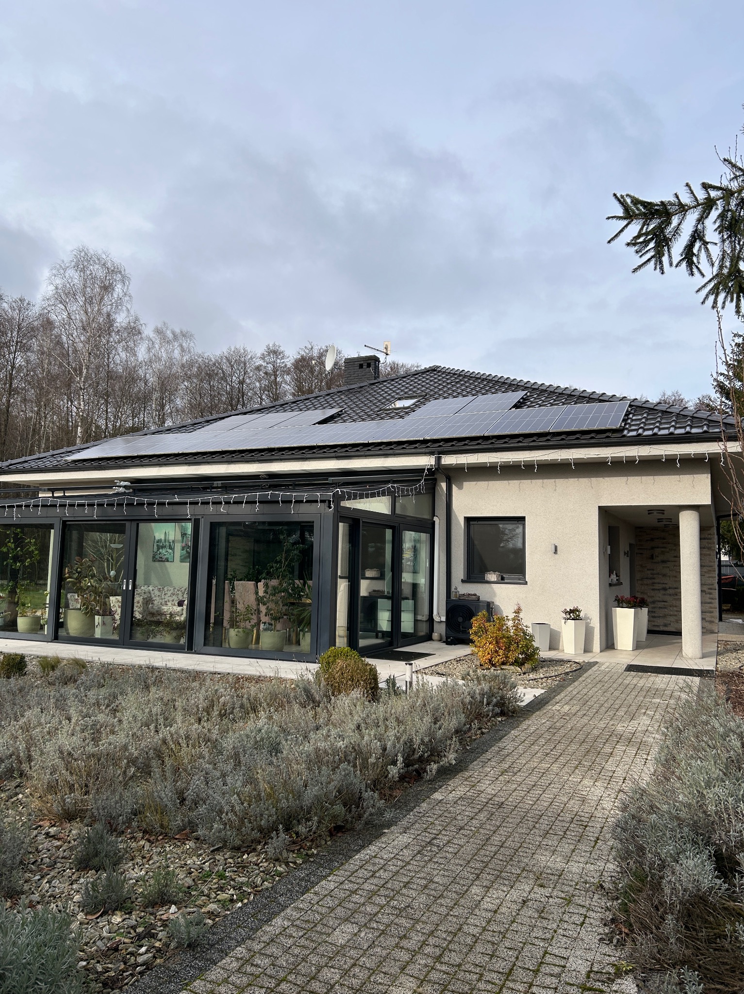 Dom jednorodzinny z panelami fotowoltaicznymi na dachu, widok z ogrodu. Szara elewacja, przeszklona oranżeria, kostka brukowa i roślinność na pierwszym planie.