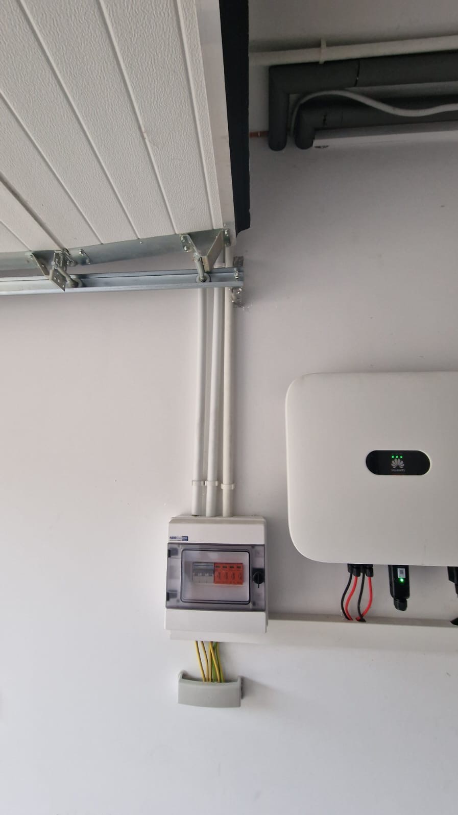 Biała ściana z zamontowaną skrzynką elektryczną, przewodami i falownikiem Huawei z zielonymi diodami. Widoczne rury instalacyjne i fragment bramy garażowej.