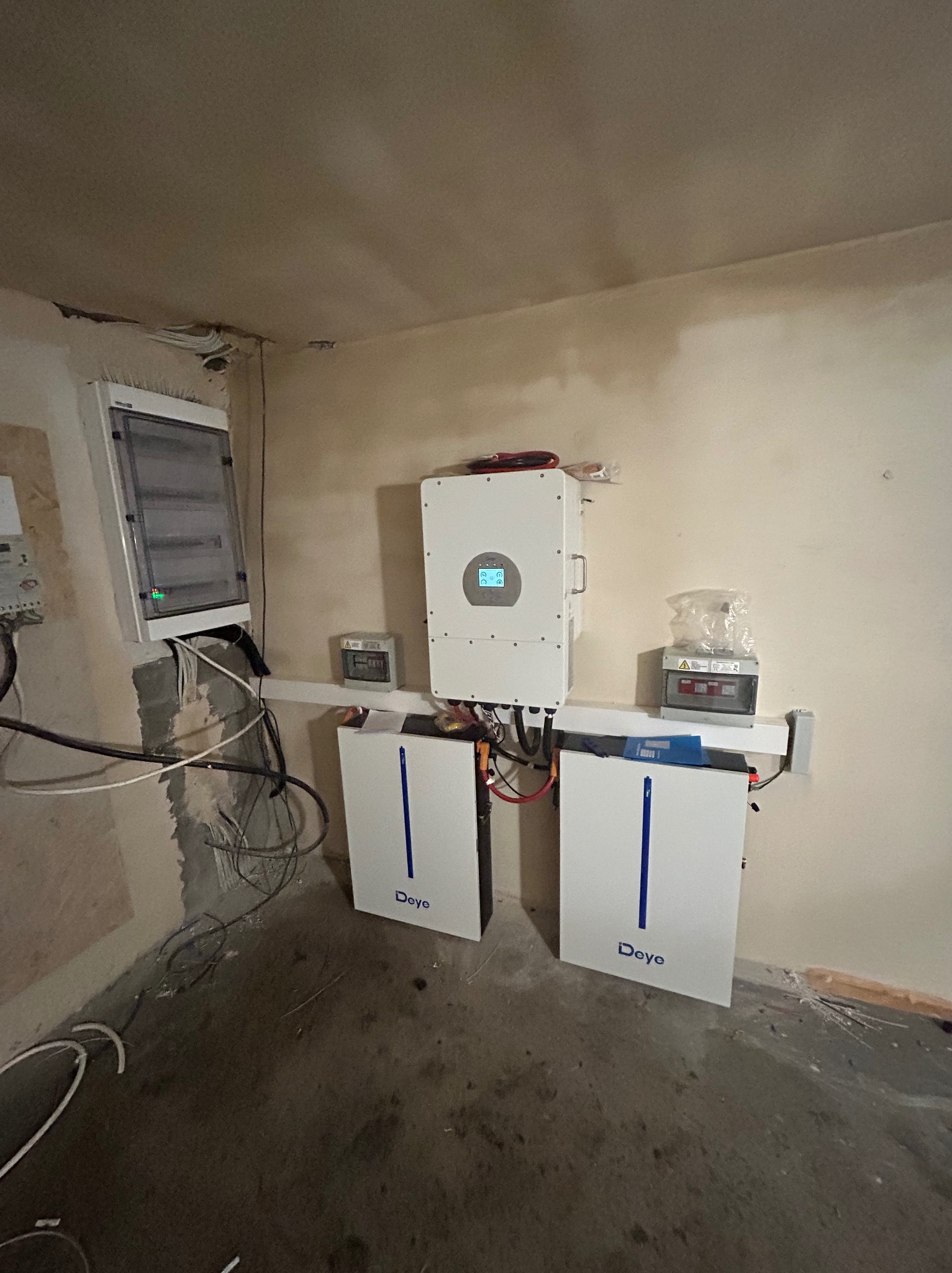 Białe magazyny energii Deye z inwerterem i rozdzielnicą na ścianie w surowym pomieszczeniu. Widoczne kable i instalacje elektryczne.