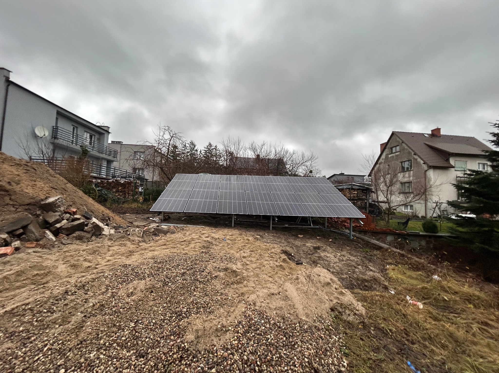 Panel fotowoltaiczny na stelażu, widok z dołu na tle pochmurnego nieba i zabudowań. Teren nierówny, z gruzem i ziemią. Instalacja w trakcie prac ziemnych.
