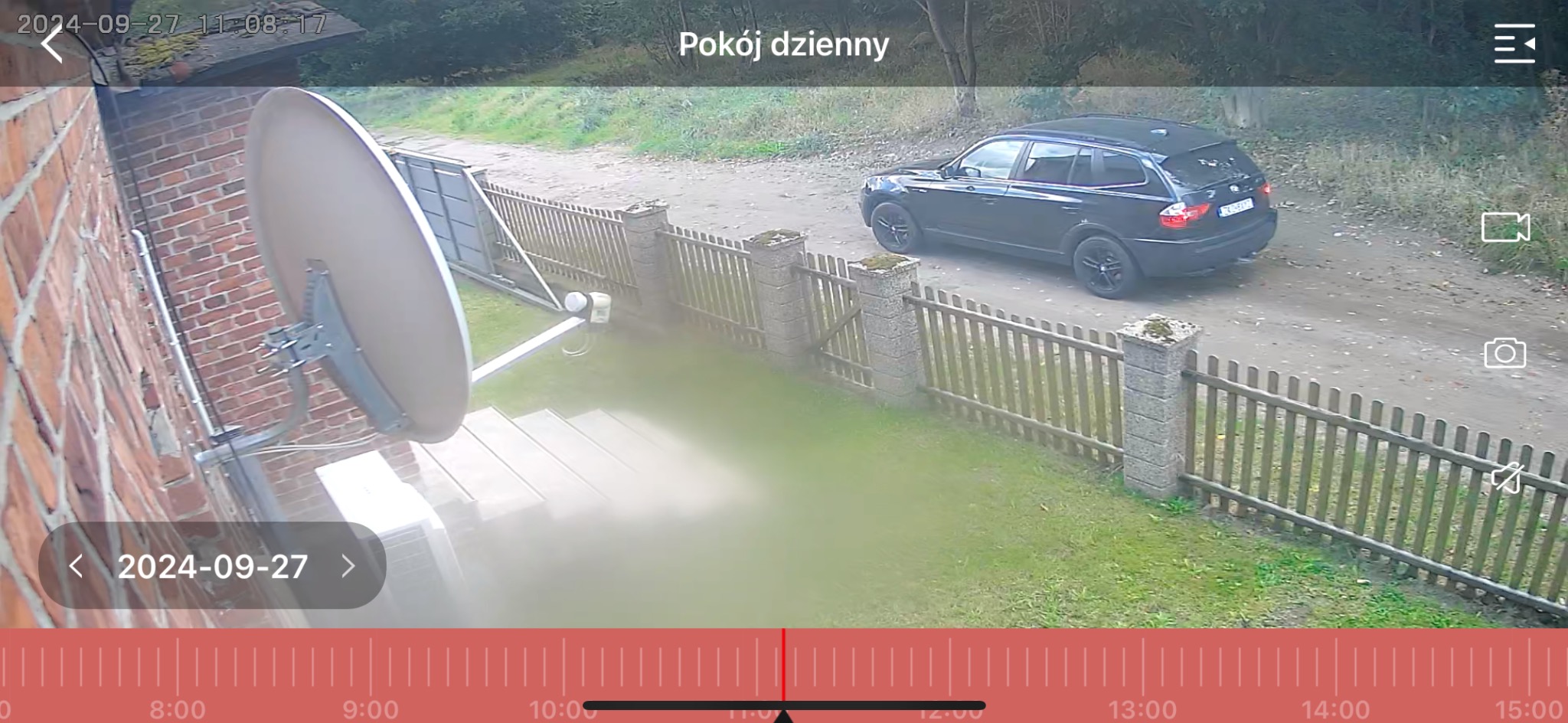 Zewnętrzny monitoring domu: antena satelitarna na ceglanej ścianie, widok na podjazd z czarnym autem i drewnianym ogrodzeniem. Zrzut ekranu z aplikacji monitoringu.